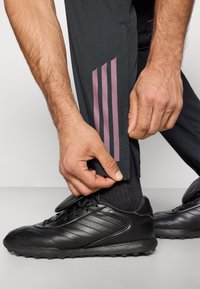 Αθλητικό παντελόνι σε σκουρόγκρι ύφασμα με ροζ τριπλή ρίγα. Ένα χέρι ρυθμίζει το μανσέτο του παντελονιού, αποκαλύπτοντας μαύρα αθλητικά παπούτσια με υφή στην επιφάνεια.