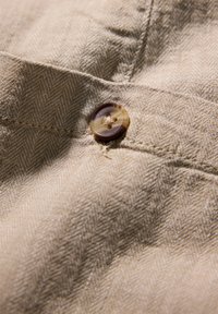 Beige stof met een visgraatpatroon, met een bruin gemarmerde knop erop genaaid. Gestructureerd en lichtgewicht materiaal.