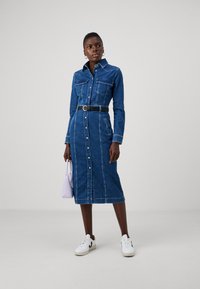 Jeansskjol med långa ärmar, knappknäppning fram, två bröstfickor och ett figursytt midjeband, tillsammans med vita sneakers och en ljuslila väska.