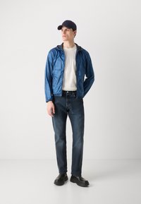 Blaue leichte Jacke mit Kapuze über einem weißen T-Shirt, kombiniert mit dunklen Jeans und schwarzen Schuhen. Das Model trägt eine marineblaue Mütze.