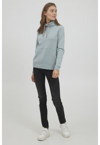 Felpa azzurra con collo a imbuto, tasca a marsupio, cordini a contrasto e polsini a coste, abbinata a jeans neri slim-fit e sneakers bianche.