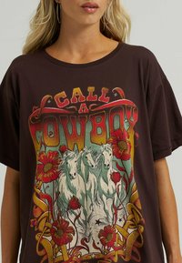 Brązowy, oversize t-shirt z wielokolorową grafiką przedstawiającą trzy białe konie otoczone czerwonymi kwiatami oraz napisem "ZADZWOŃ DO KOWBOJA".
