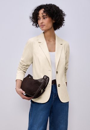 Femme aux cheveux bouclés portant un blazer crème et un jean bleu, tenant un sac à main tissé marron foncé sur un fond gris clair.