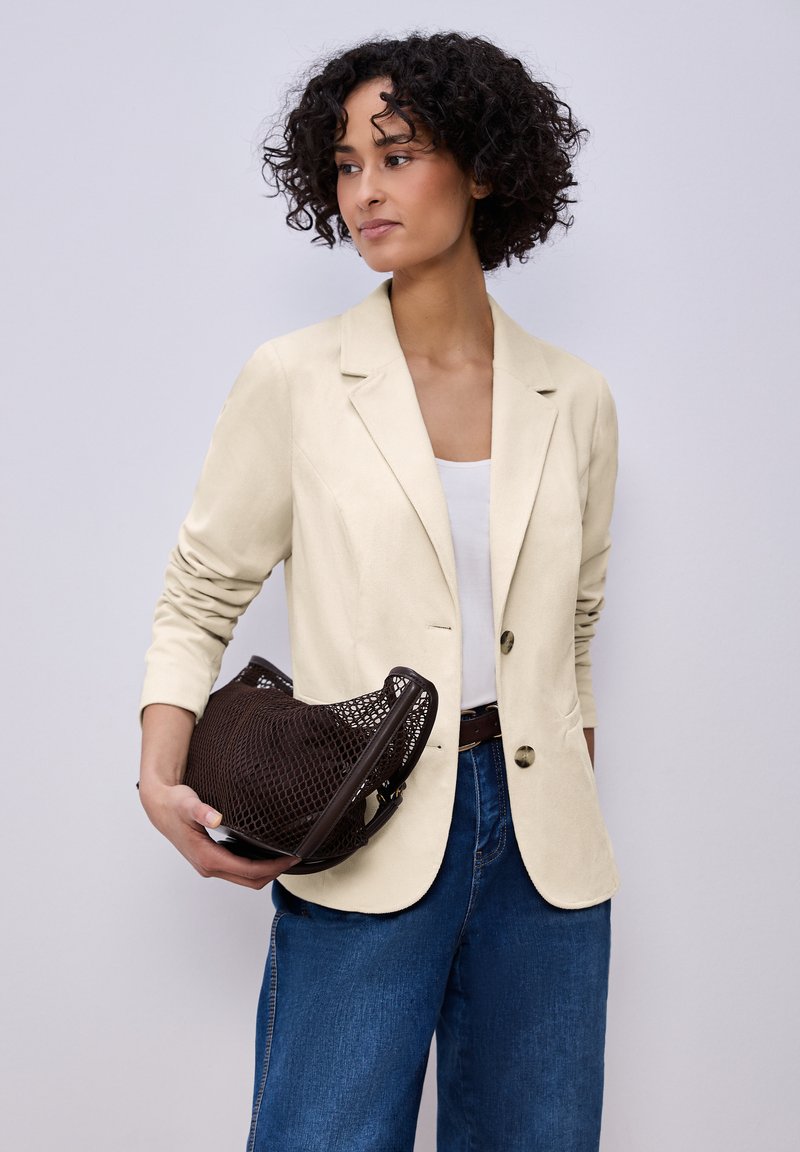 Femme aux cheveux bouclés portant un blazer crème et un jean bleu, tenant un sac à main tissé marron foncé sur un fond gris clair.