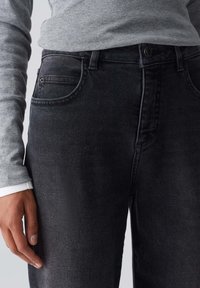 Jeans de mezclilla negra con corte Slim, con un bolsillo frontal, un ligero desgastado y un cierre clásico de botón. Camisa de manga larga gris por debajo.