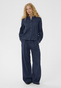 Göndör szőke hajú nő sötétkék farmeringben és bő szárú nadrágban, zsebeibe tett kézzel, fehér háttér előtt áll.