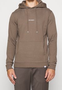 Hombre con sudadera con capucha marrón y bolsillo frontal, con el texto blanco "LES DEUX" centrado en el pecho.