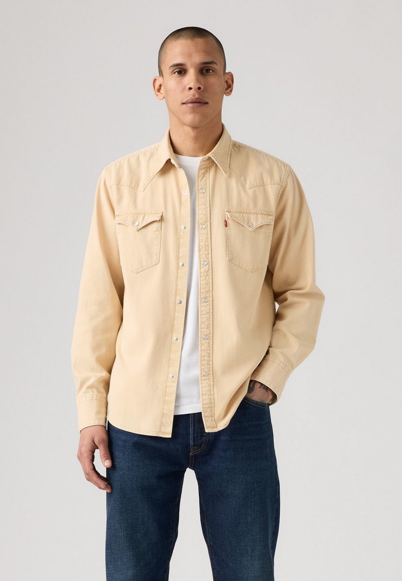 Mann iført en beige skjorte med knapper over en hvit t-skjorte og mørkeblå jeans, står med den ene hånden i lommen mot en ensfarget bakgrunn.