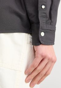 Camicia grigio scuro con colletto, dalla texture liscia, con bottoni bianchi sul polsino e orlo dritto, abbinata a pantaloni di colore chiaro.