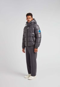 Puffjakke i mørkegrå med hætte, med en blå detalje på ærmet og en lynlåslukning foran. Bæres med grå bukser og hvide sneakers.