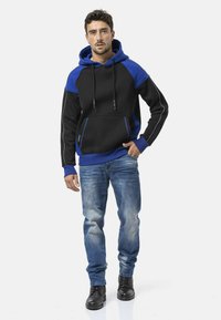 Sweat à capuche noir et bleu avec une poche avant, présentant des manches contrastées, des cordons de serrage et un liseré réfléchissant. Porté avec un jean bleu clair et des bottes noires.