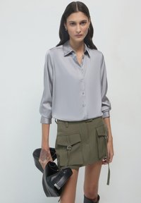 Camicia button-up in satin grigio chiaro con maniche lunghe e colletto, abbinata a una gonna cargo verde oliva e una borsa nera a tracolla.