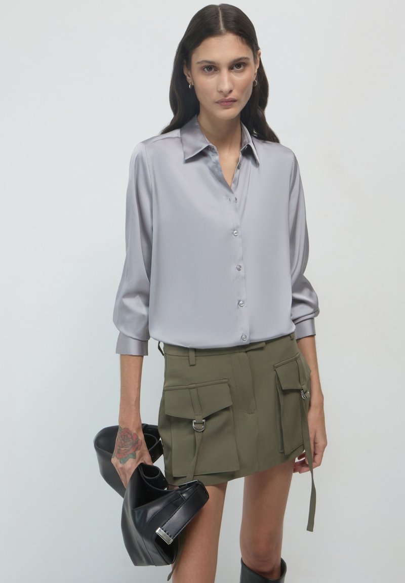 Camicia button-up in satin grigio chiaro con maniche lunghe e colletto, abbinata a una gonna cargo verde oliva e una borsa nera a tracolla.