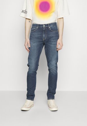 Vaqueros de mezclilla azul con un corte ajustado, que presentan una cintura de altura media, un diseño de cinco bolsillos y piernas ligeramente desteñidas, combinados con una camiseta de textura color crema.
