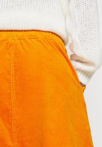 Manche de pull en maille blanche et jupe en velours côtelé orange avec taille élastique, main reposant dans la poche de la jupe.