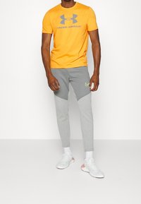 Oranžové tričko s krátkým rukávem s šedým logem "Under Armour", které je kombinováno se šedými zúženými tepláky s tmavě šedým panelem a neonově žlutým logem Nike.