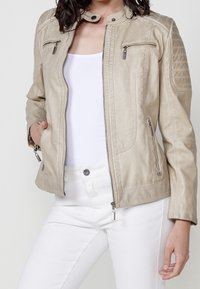 Beżowa kurtka z ekoskóry z kołnierzem stójką, zapinana na zamek, z dwoma kieszeniami na piersi oraz pikowanymi detalami na ramionach. Gładka tekstura, dopasowany krój.