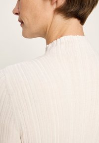 Profil einer Person mit kurzen braunen Haaren, die einen cremefarbenen gerippten Pullover mit einem hohen, gewellten Kragen trägt, vor einem schlichten Hintergrund.