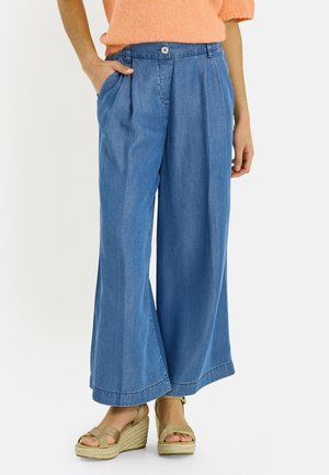 Mujer con pantalones vaqueros de pierna ancha en azul, un suéter naranja y sandalias de cuña beige, con una mano en el bolsillo.