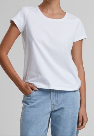 Femme portant un t-shirt blanc uni à manches courtes et un jean bleu clair, une main dans une poche, sur un fond uni.
