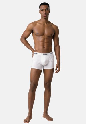 Muskeløs mand, der står barfodet iført hvide Calvin Klein boxer shorts mod en ensfarvet lys baggrund.
