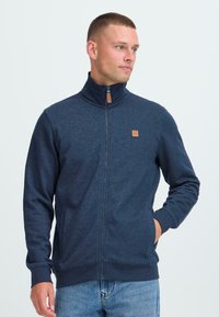 Muž s krátkymi vlasmi, ktorý má na sebe tmavomodrú zipsovú bundu s koženou nášivkou, pozerajúci doprava a s jednou rukou v vrecku.