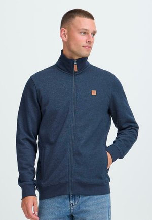 Man med kort hår som bär en marinblå zip-up jacka med en läderlogotyppatch, tittar åt höger med en hand i fickan.