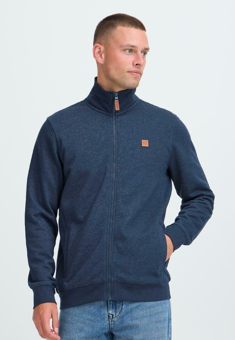 Muž s krátkymi vlasmi, ktorý má na sebe tmavomodrú zipsovú bundu s koženou nášivkou, pozerajúci doprava a s jednou rukou v vrecku.