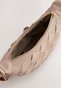 Beige vävd axelremsväska med en slät yta, prydd med en gyllene dragkedja och ett tygfoder inuti. Accenterad med en robust rem.