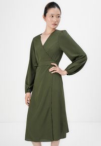 Olive green wrap dress with long puff sleeves, V-neckline, and a side slit. Made of smooth fabric with a fitted waist. Simple design details.