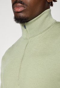 Lys grønn ribbestrikket halv-zip genser med høy krage og strukturert stoff, med en kontrasterende glidelås i en mørkere nyanse.