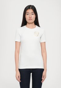 T-shirt branco de manga curta feito de algodão suave com um decote redondo, apresentando um logotipo redondo dourado na área do peito esquerdo.