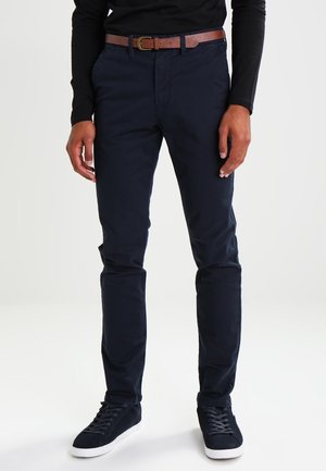 Mann trägt marineblaue Slim-Fit-Hose, braunen Ledergürtel, schwarzes Langarmshirt und marineblaue Sneakers mit weißen Sohlen, steht auf weißem Boden.