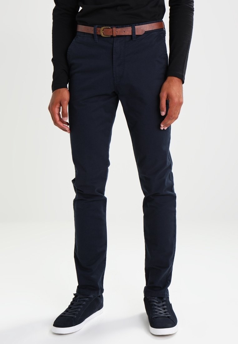 Homme portant un pantalon slim bleu marine, une ceinture en cuir marron, une chemise noire à manches longues et des baskets bleu marine avec des semelles blanches, debout sur un sol blanc.