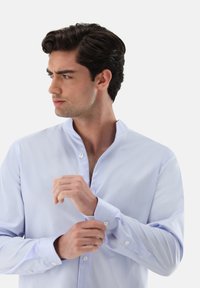 Hellblaues Langarmhemd mit Stehkragen, Ein-Knopf-Manschetten und glatter Textur. Auffälliges Knopfdetail an den Manschetten und im Kragenbereich.