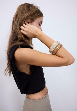 Ung kvinde med langt brunt hår iført sort ærmeløs top og beige bukser justerer sit hår med venstre hånd, beige armbånd med nitter på håndleddet.