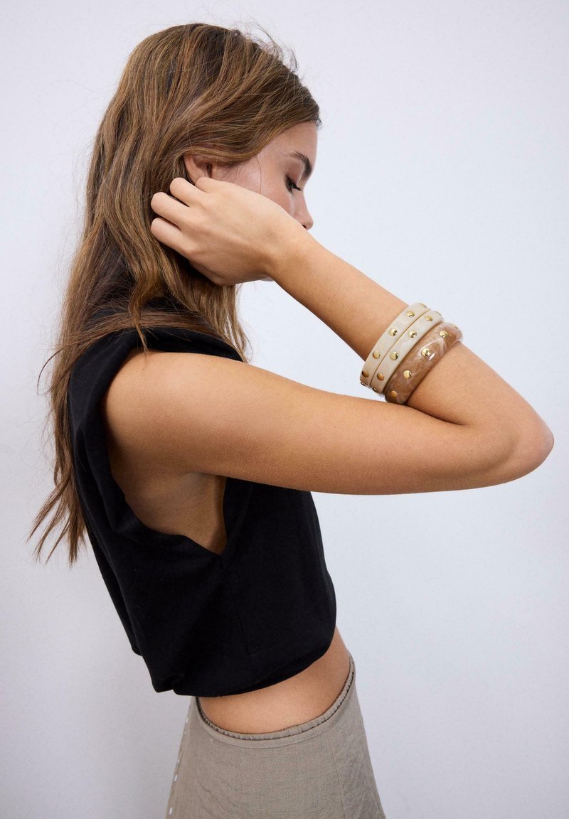 Ung kvinde med langt brunt hår iført sort ærmeløs top og beige bukser justerer sit hår med venstre hånd, beige armbånd med nitter på håndleddet.