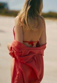 Roter, transparenter Überwurf über einem rosa Bikini, mit einem bindbaren Design und strukturiertem Stoff. Weiche Wellen im Hintergrund, sichtbarer Sandstrand.