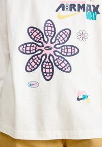 Hvit t-skjorte med et stort rosa blomstermotiv med rutenett-lignende kronblader, flere Nike-logoer og tekstuliknelse i svart og teal.