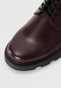 Bota de cuero burdeos con un acabado suave y brillante, cordones negros y una suela de goma duradera. Puntera redondeada y detalles de costura minimalista.