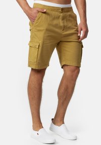 Mand iført sennepsfarvede cargo shorts med sidelommer og hvide slip-on sko, stående med den ene hånd i lommen.