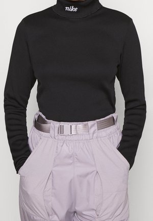 Personne portant un col roulé noir à manches longues avec le logo blanc Nike et un pantalon taille haute violet clair avec une large ceinture et les mains dans les poches.