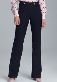 Kobieta w czarnych, wysokich, prostych spodniach, różowej bluzce w kropki i jasnoróżowych czółenkach, stojąca na szarym tle.