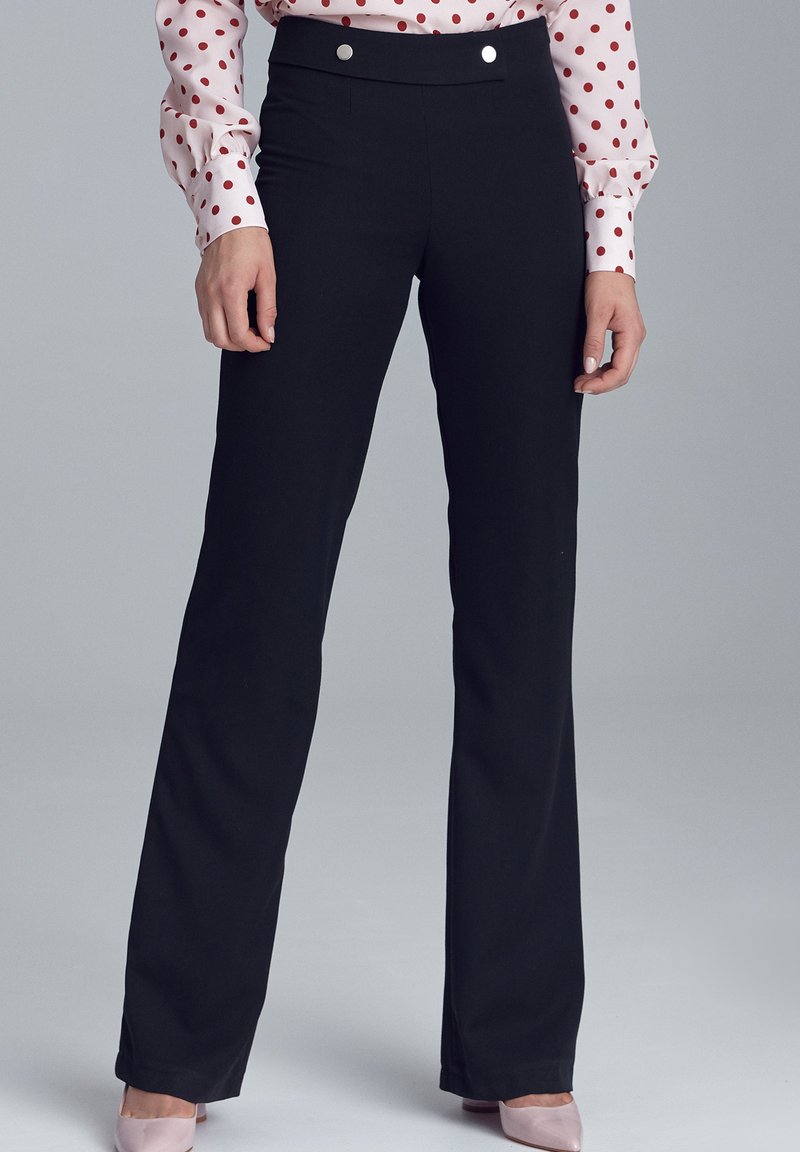 Kobieta w czarnych, wysokich, prostych spodniach, różowej bluzce w kropki i jasnoróżowych czółenkach, stojąca na szarym tle.