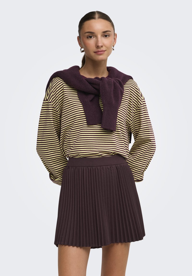 Jeune femme avec les cheveux attachés, portant des boucles d'oreilles créoles, une chemise à manches longues rayée jaune et marron, un pull violet sur les épaules et une jupe plissée marron.