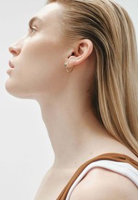 Pendiente de aro de oro con un pequeño acento de turquesa, llevado en una oreja perforada. Se puede ver cabello rubio largo y liso y una piel suave.