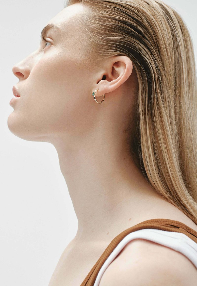 Pendiente de aro de oro con un pequeño acento de turquesa, llevado en una oreja perforada. Se puede ver cabello rubio largo y liso y una piel suave.