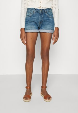 Shorts en denim avec des revers roulés, délavage bleu clair et design à cinq poches. Associés à des sandales à lanières beiges sur pieds nus.