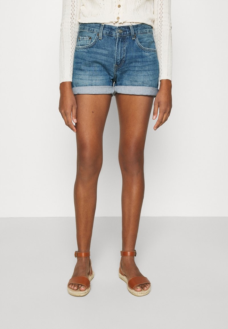 Denimshortsit, joissa on käännetyt hapsut, vaalea sininen pesu ja viisi taskua. Yhdistetty ruskeisiin hihnasandaaleihin paljain jaloin.