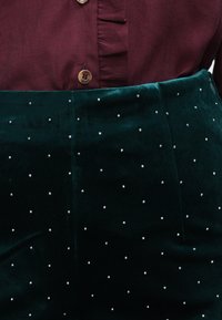 Smaragdrohelised sametpüksid, mida ehtivad hõbedased nõelad, millel on sile tekstuur ja keha lähedane lõige, koos marooni värvi nööbiga särk.